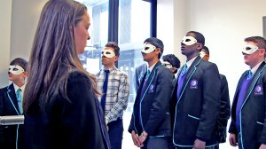 Seven teenage boys are standing together at the bottom of a school stairway, looking up, wearing white masks covering their eyes. A girl without a mask is standing across from them, looking toward them.