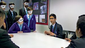 In a classroom, nine students are grouped around a long, rectangular table talking to each other. 