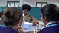 The backs of two sitting students are facing a male teacher who is standing, smiling, and looking proudly at a booklet he's holding.