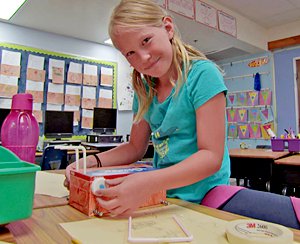 Girl at a table putting water bottle top wheels on a tissue box