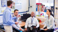 Four teachers are sitting in the corner of an office, smiling, laughing, and talking to each other. 