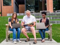 Three adults sitting side-by-side on a bench working on their laptops