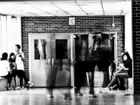 Three students are sitting on a large window sill on the left side of a high school hallway. Two students are sitting on a window sill on the right side of the school hallway. In the middle of the hallway is a faint, blurred image of three teenage student