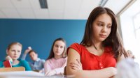 A girl looks lonely in class as two peers look angry behind her.