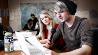 Boy reading from a piece of paper in front of a laptop; girl sitting next to him looking on