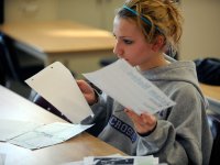 A teenage girl in a gray sweatshirt is sitting at her paper-scattered desk, holding pieces of paper in both hands.