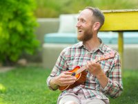 Bearded man sitting outside, smiling, playing the ukulele