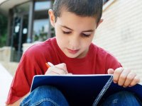 A young boy in a red shirt is sitting outside with a building behind him. His knees are up close to his chest with a with a blue notebook resting against them. He's smiling, writing in his notebook.