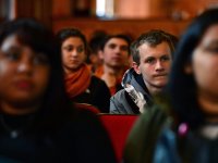 High school students are sitting in red chairs in a theatre. A teenage boy, two rows back from the front, is in focus; everyone else is slightly blurry.