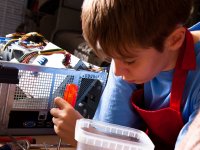 A closeup of a young boy in a blue shirt and red apron leaning over a table with a screw driver in his hand, a box of electronics next to him.