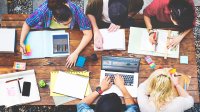 An aerial shot of five high school students sitting at a wooden picnic table outside with laptops, tablets, and notebooks open, working on math problems with rulers and graphs.