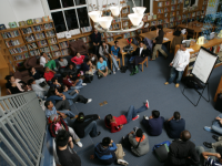 a photo of a group of high school students sitting on the floor in a discussion with their teacher.