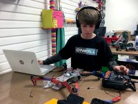 A young boy wearing a black, long-sleeve shirt and headphones has one arm extended next to his laptop, and the other hand is touching a remote control. In between his laptop and the remote control is a robotic device made out of plastic, metal, and wires.