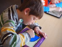 A boy is hunched over a rectangular device on his desk, drawing.