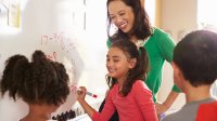 A group of young students work on a math problem on a whiteboard as the teacher watches.