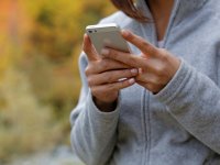 A closeup of a young woman's hands holding an iPhone. She's wearing a grey, full zip sweatshirt, and the autumn-colored background is blurred out.