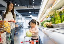 A mom and daughter are walking side by side in the produce aisle of a grocery store, pushing a regular and kid-sized shopping cart.