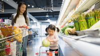 A mom and daughter are walking side by side in the produce aisle of a grocery store, pushing a regular and kid-sized shopping cart.