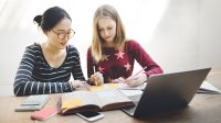 Two girls look at a book and write on Post-its.