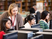 A librarian is peering over the shoulder of a student who is sitting next to two other female students, all working on desktop computers in a library.