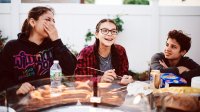 Two teenage girls are sitting outside at a glass table, eating lunch and laughing.