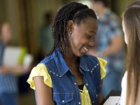 A teenage girl in a yellow shirt and jean vest is smiling, looking down at an opened binder in her hands, showing it to another girl outside of the frame of the photo. The rest of the photo is blurred out, and seems to be students in a school hallway.