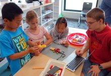 Four young students are sitting around a classroom table building a contraption out of legos and wires. It looks like it might be able to move prompted by technology.