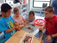 Four young students are sitting around a classroom table building a contraption out of legos and wires. It looks like it might be able to move prompted by technology.