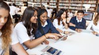 Five teenaged students are seated at a table looking at computer screens, talking, and smiling. 