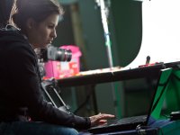 A female teacher is sitting, wearing a hoodie and jeans, looking down at and typing on a laptop that's near her knees on a short table. She's in a classroom with a camera, a projector, and a light on a poll behind her.