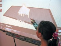 A closeup of a young girl standing next to and pointing at a projector screen. A 3D drawing made up of small squares is projected on the screen, showing the inside of a white room with a brown floor and a small white pyramid in the middle.