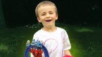 A young boy standing outside in the grass has a robotic right hand, and he's smiling, holding a blue and red plastic horseshoe.