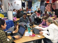 Three groups of four to five teachers are sitting at tables in a classroom talking to each other, looking at their laptops, and writing on paper.