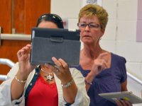 Two adult women are standing in a classroom with tablets in their hands. One of them is holding up their tablet, and both women are looking at it.