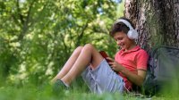 Elementary-aged boy reading a book outside while wearing headphones
