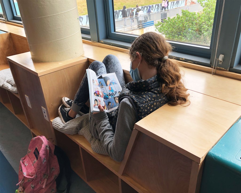Photo of a female student reading with a mask on in a school library nook by the window