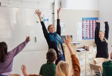 Photo of school teacher and students raising both hands in classroom