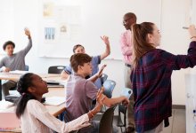 A photo of a classroom where students are raising their hands to ask a question to a fellow student who is writing something on a whiteboard. The teacher is looking over the classroom with a smile on his face.