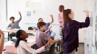 A photo of a classroom where students are raising their hands to ask a question to a fellow student who is writing something on a whiteboard. The teacher is looking over the classroom with a smile on his face.