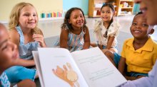 An image of elementary students sitting in a circle and looking engaged as their teacher reads them a picture book out loud.
