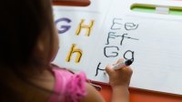 A photo of a young elementary student practicing writing their letters using a dry-erase marker.