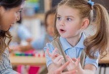 A photo of a preschool student counting on her fingers with her teacher beside her. Behind her are two other students counting blocks.