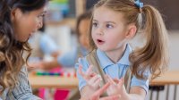 A photo of a preschool student counting on her fingers with her teacher beside her. Behind her are two other students counting blocks.