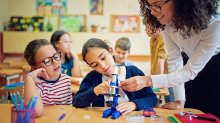 A photo of a teacher guiding an elementary student to use a microscope as the student's desk partner watches.