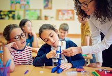 A photo of a teacher guiding an elementary student to use a microscope as the student's desk partner watches.