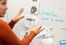 A photo of a teacher gesturing towards images of kitchen utensils taped on a white board. "Utensilios para la cocina" is written on the white board.