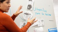 A photo of a teacher gesturing towards images of kitchen utensils taped on a white board. "Utensilios para la cocina" is written on the white board.