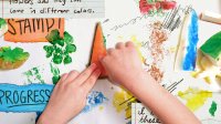 A student's hands pressing down on a carrot painted with green acrylic paint to make a print. Scattered around the hands are various stamps made from vegetables, scraps of paper, and pen doodles.
