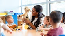 A photo of an elementary teacher sitting at a table with her students as she uses a puppy doll to engage them in a lesson.