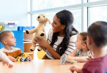 A photo of an elementary teacher sitting at a table with her students as she uses a puppy doll to engage them in a lesson.